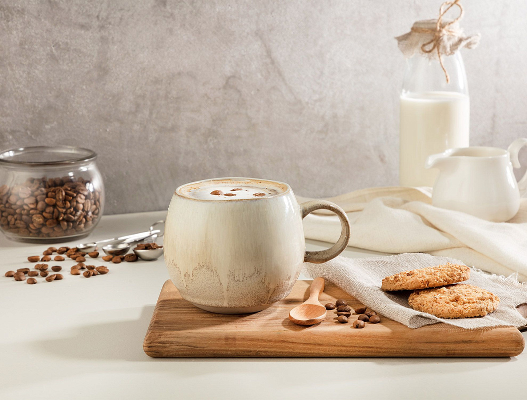 Frothy latte in a speckled ceramic mug with coffee beans, oatmeal cookies, and a wooden spoon on a rustic board beside a jar of coffee beans and a milk bottle